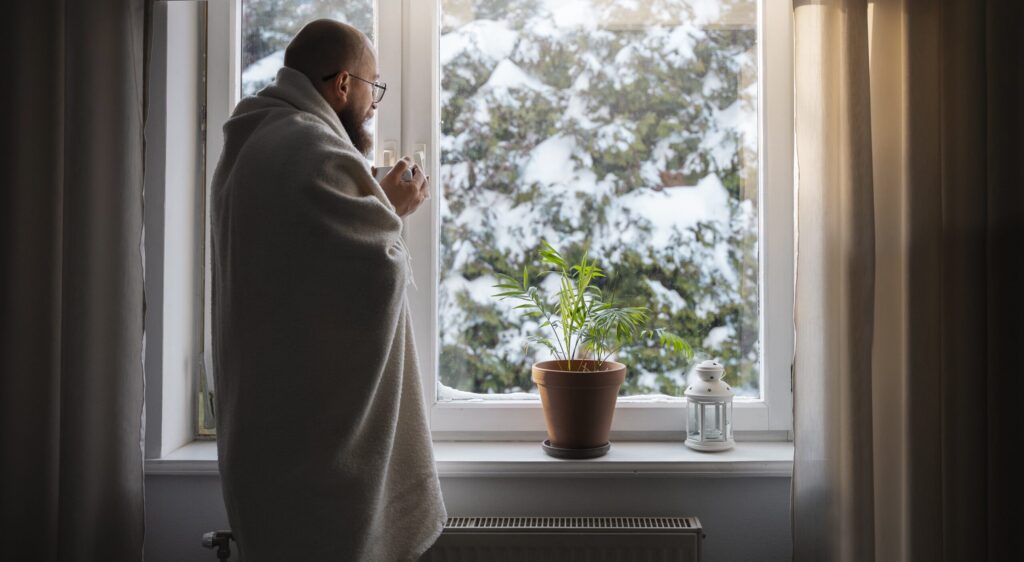 Man staat met deken om voor het raam. Buiten ligt sneeuw.