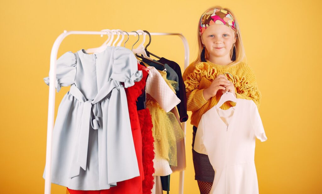 Blond meisje staat naast een kledingrek met kinderkleding.