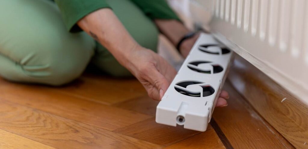 Vrouw zit op haar knieen met een radiator ventilator in haar handen en plaatst die net boven de vloer onder de radiator.