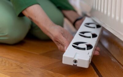 Vrouw zit op haar knieen met een radiator ventilator in haar handen en plaatst die net boven de vloer onder de radiator.