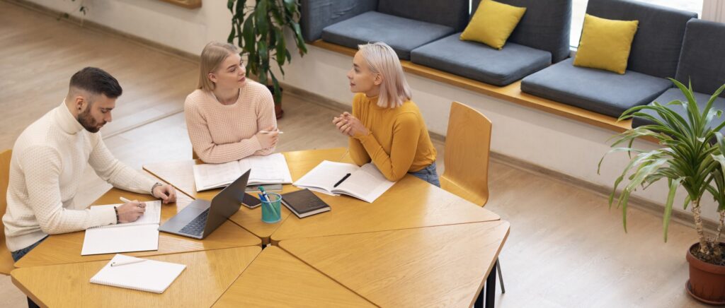 Drie mensen vergaderen aan tafel, met laptop en notities.