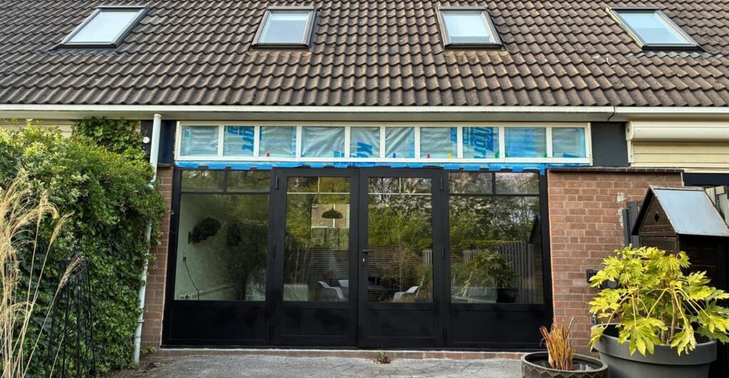 Gezien vanuit de tuin: net geplaatste zwarte kozijnen van openslaande deuren. Daarboven is nog isolatiemateriaal zichtbaar.