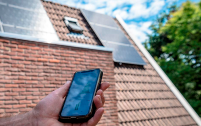 Zonnepanelen op een schuin dak. Een hand houdt een smartphone vast met daarop de app van de energieleverancier.