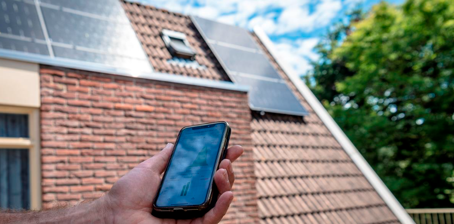 Zonnepanelen op een schuin dak. Een hand houdt een smartphone vast met daarop de app van de energieleverancier.