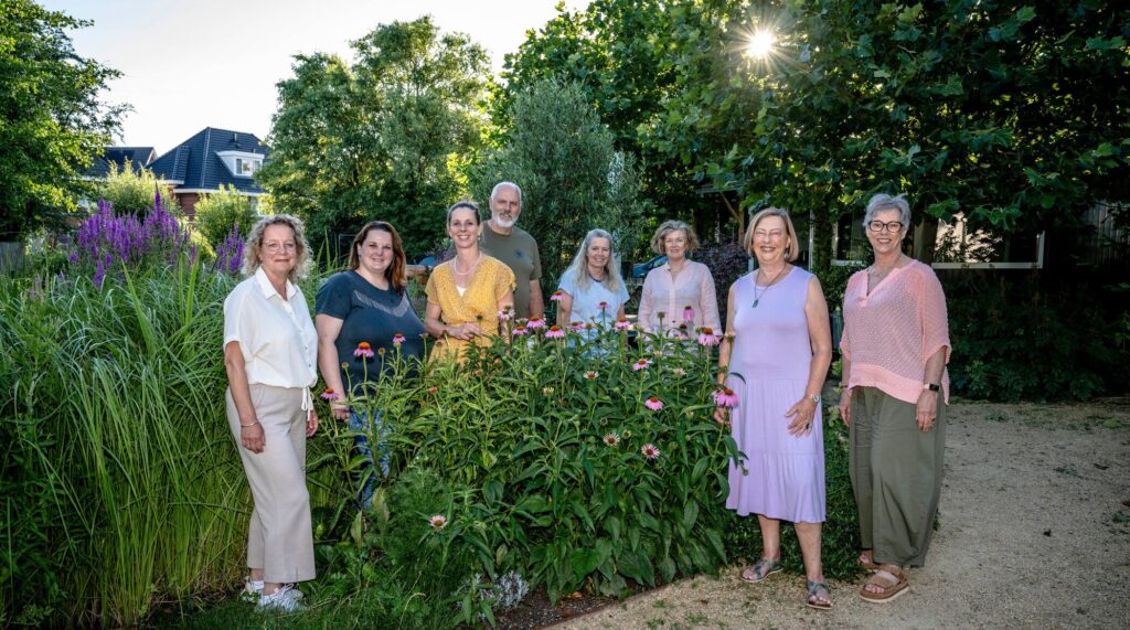 Groepsfoto van 8 mensen in het groen. Zomerse kleding, zon schijnt door de bomen.